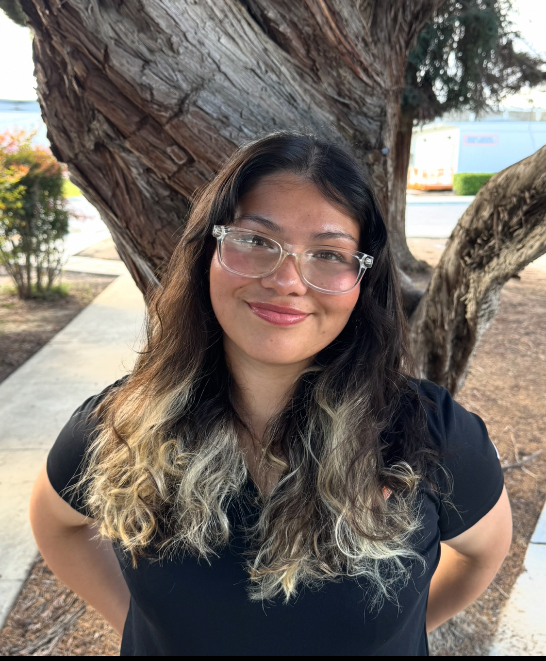A smiling woman with long, wavy hair stands confidently in front of a tree. She wears clear glasses and a black shirt.
