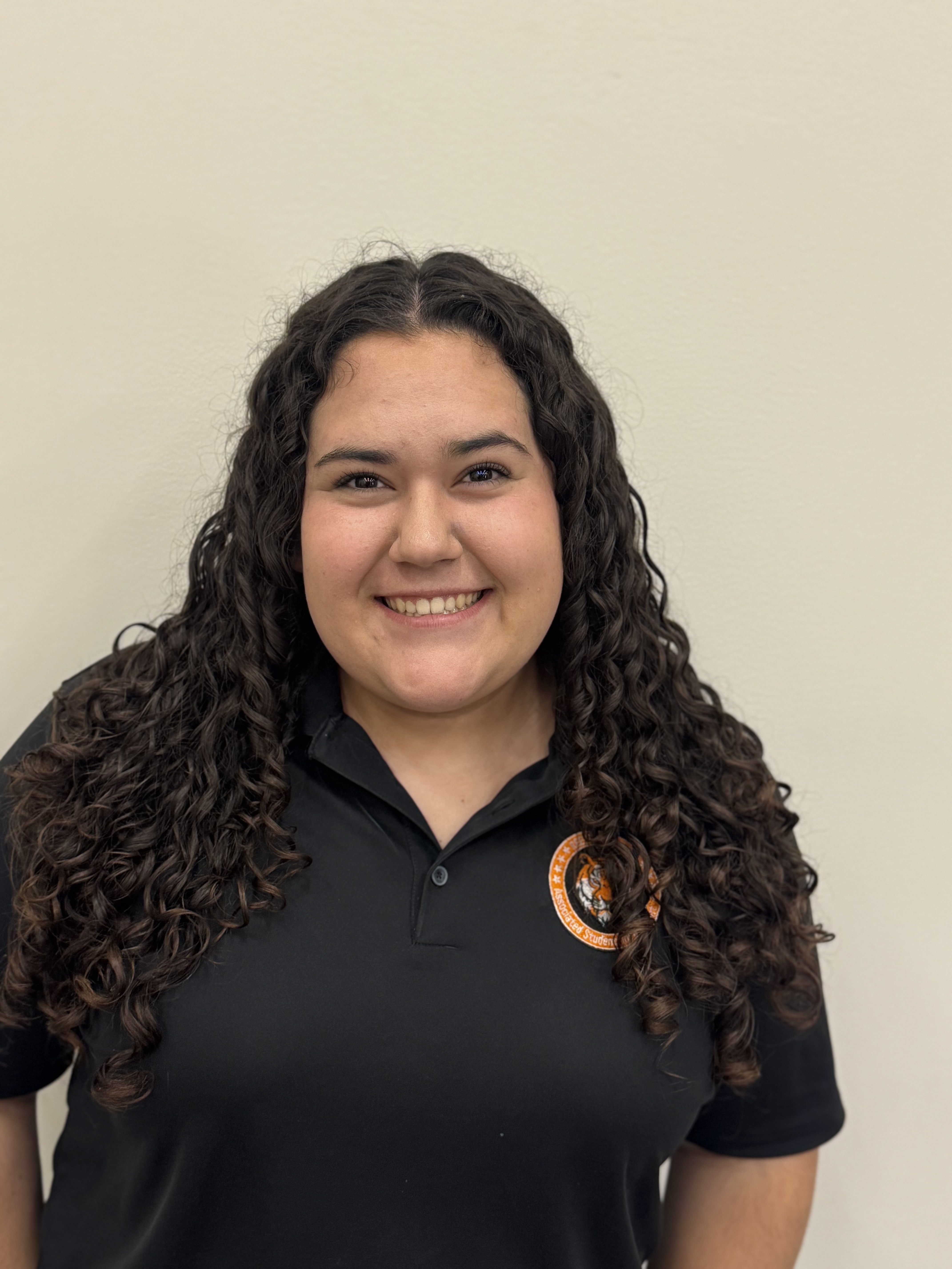 Portrait of a smiling young woman with long, curly hair, wearing a black polo shirt with an orange logo.